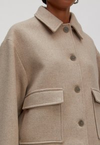 Close-up van een beige wollen jas met grote kn openingen en klepzakken, gedragen door een persoon met een ringoorbel en een gebruinde huid.