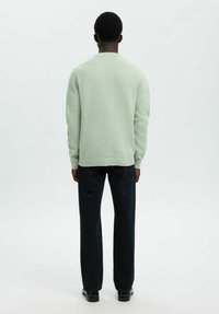 Mężczyzna w jasnoszarej bluzie, czarnych dżinsach i czarnych butach, stojący plecami do obiektywu na tle białej ściany.