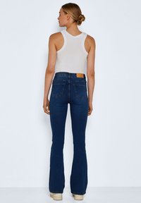Blue flared jeans with a high waist, back pockets, and a tan leather patch at the waistband. Paired with a fitted white tank top.