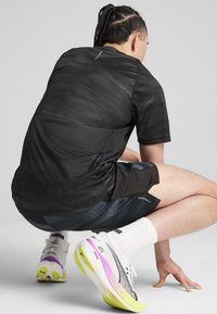 Sort atletisk skjorte med et struktureret mønster, parret med mørke shorts. Hvide og multicolore løbesko med et karakteristisk såledesign.