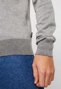 Gray knit sweater with ribbed cuffs and hem, featuring a small black brand label on the side. Paired with blue denim pants.