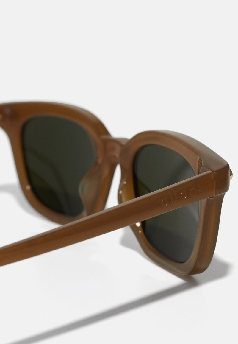 Occhiali da sole in plastica marrone con lenti verdi, design angolare e logo inciso sulla cerniera, con una texture liscia e forma classica.