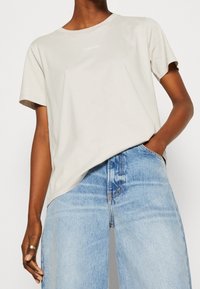 T-shirt in cotone beige chiaro con colletto girocollo e maniche corte; presenta un design semplice e un piccolo logo bianco "Calvin Klein" sul petto.