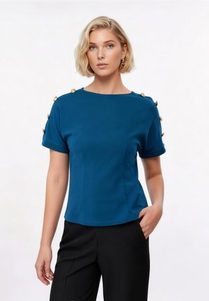 Γυναίκα με κοντά ξανθά μαλλιά, φοράει μπλε-πράσινο μπλουζάκι με κοντά μανίκια και χρυσά κουμπιά στους ώμους και μαύρο παντελόνι, στέκεται με το ένα χέρι στην τσέπη.