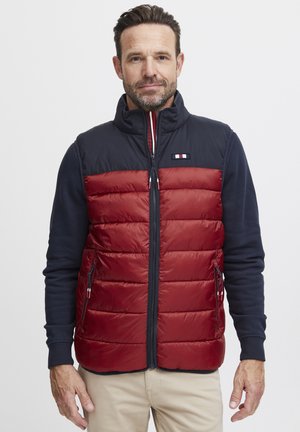 Red puffer vest with navy shoulders, zippered front, and side pockets. Features a logo patch and a smooth, quilted texture.