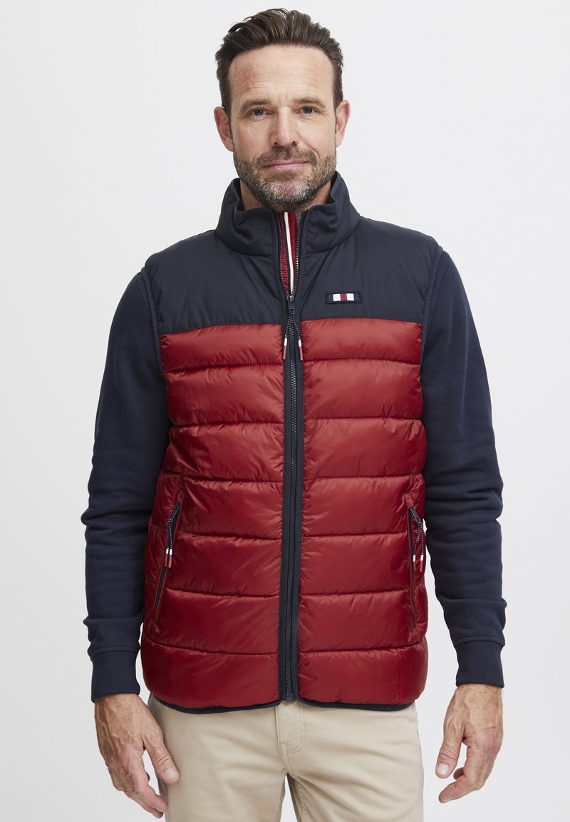 Gilet rembourré rouge avec épaules navy, devant zippé et poches latérales. Présente un patch logo et une texture lisse et matelassée.
