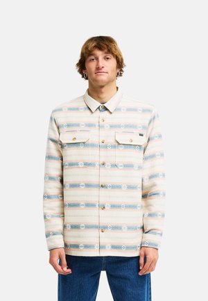 Junger Mann mit hellbraunen Haaren, der ein beigefarbenes und blau gestreiftes Hemd mit Knöpfen und blaue Jeans trägt, steht vor einem einfarbigen Hintergrund.