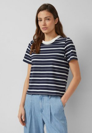 Jonge vrouw die een marineblauw-wit gestreept shirt met korte mouwen en lichtblauwe, hooggesneden spijkerbroek draagt, staande voor een effen achtergrond.