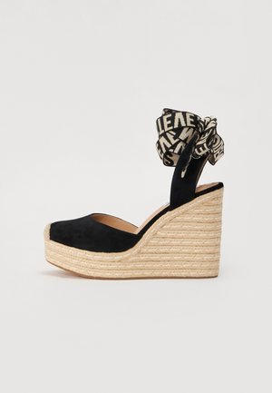 Scarpa con zeppa in suede nero con suola in iuta intrecciata. Presenta una fascia decorativa in tessuto con un motivo contrastante annodata intorno alla caviglia.