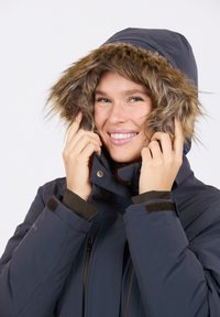 Marineblaue Parka mit einer mit Fell gefütterten Kapuze, ausgestattet mit einem Reißverschluss, verstellbaren Bündchen und glattem Stoff. Das Modell zeigt den Hoodie-Stil.