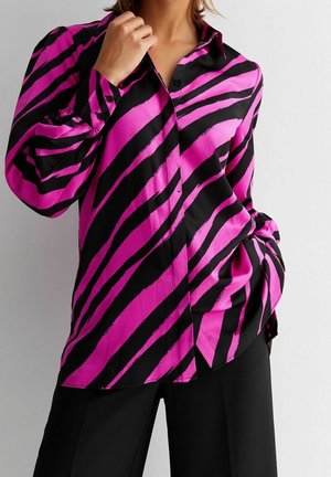 Femme portant une chemise à manches longues boutonnée avec de larges rayures diagonales noires et roses zébrées, associée à un pantalon noir.