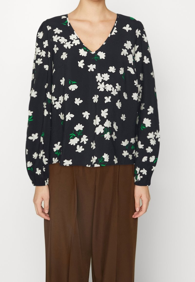 Blomsterbluse i mørk navy med hvide blomster og grønne blade, med V-hals og lange, samlede ærmer. Båret med brune bukser.
