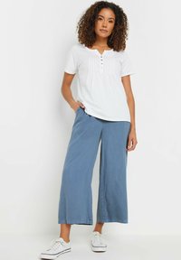 Witte kortemouwen shirt met plooien en knoopdetails, gecombineerd met losse blauwe wijde broek en witte sneakers.