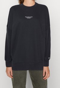 Persona che indossa una felpa nera a maniche lunghe con il testo bianco "Abercrombie and Fitch" in piccolo sul petto e "A&F" sul polso sinistro.