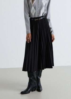 Donna con cardigan grigio a rombi, gonna midi nera plissettata con cintura e stivali in pelle neri con tacco, in piedi su un pavimento chiaro.