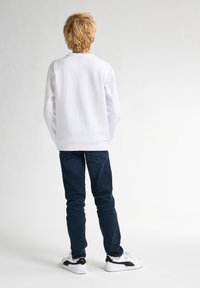 Witte sweatshirt met lange mouwen en een relaxte pasvorm, gecombineerd met donkerblauwe jeans. De outfit wordt aangevuld met witte sneakers met zwarte accenten.