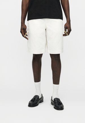 Mies, jolla on valkoiset polven mittaiset shortsit, mustat hapsulliset loafersit, valkoiset puuvillasukat ja musta paita, seisomassa valkoista taustaa vasten.