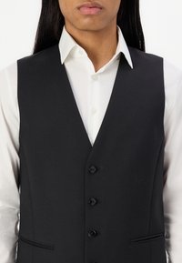 Schwarze Weste mit Reverskragen, fünf Knöpfen und slim fit. Getragen über einem weißen Hemd mit glatter Textur und maßgeschneidertem Design.