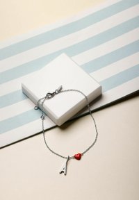 Silverkedjesarmband med en versal bokstav "A"-berlock och en liten röd hjärtberlock. Visad mot en ljus bakgrund med en låda.