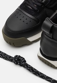 Black leather sneakers with textured detailing, rubber sole in olive green, and black laces with white stitching, featuring a pull tab at the heel.