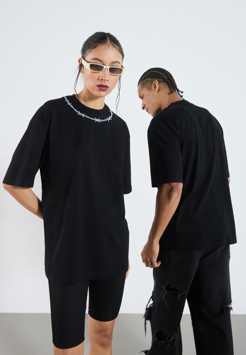 Camisetas negras de gran tamaño con bordado de alambre de púa blanco alrededor del escote. Llevadas con jeans negros y pantalones cortos ajustados de ciclismo de canalé.