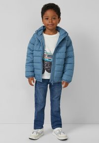 Padded blue jacket z kapturem, z zamkiem z przodu, noszony na białej koszulce z grafiką, w połączeniu z niebieskimi dżinsami i białymi trampkami.