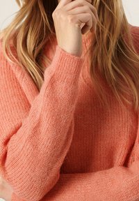 Pull en maille corail avec une coupe ample, des poignets côtelés et une texture douce. Main visible saisissant le tissu, mettant en valeur le design du col large.