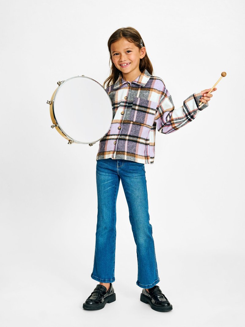 Chemise en flanelle à carreaux dans des couleurs pastel, jeans bleus à jambes évasées, chaussures à plateformes noires, tenant un tambourin et un bâton en bois.