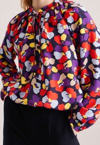 Blommig blus i livfulla färger som rött, lila, gult och blått på en mörk bakgrund. Har en rynkad halsringning och knappar framtill.