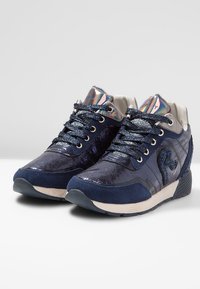 Un paio di sneaker blu navy con tessuto lucido e texturizzato, lacci glitterati, linguetta iridescente e suole bianche imbottite angolate in avanti.