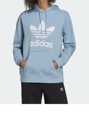 Osoba w jasnoniebieskiej bluzie z kapturem Adidas z białym logo oraz czarnych spodniach Adidas z małym białym logo, ręce w kieszeni bluzy.
