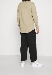 Uomo in camicia beige a maniche lunghe, pantaloni cargo neri a gamba larga, calzini bianchi e sneakers bianche Adidas in piedi su un pavimento grigio.