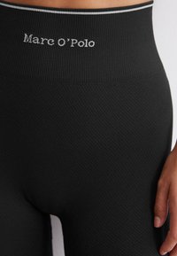 Černé legíny s texturovaným vzorem a širokým pasem, na kterém je vyšitý nápis "Marc O'Polo" v šedé barvě. Hladký povrch látky.