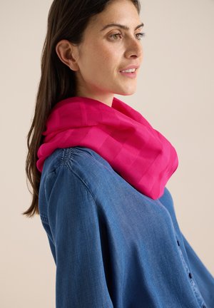 Femme aux longs cheveux bruns portant une chemise en jean bleue et une écharpe rose vif, regardant légèrement vers la droite sur un fond uni.