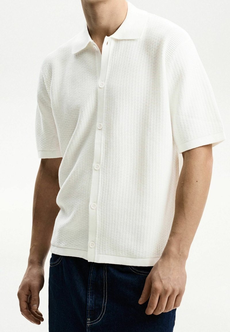 Weißes Polohemd mit kurzen Ärmeln, das ein strukturiertes Strickmuster, eine Knopfleiste und einen klassischen Kragen aufweist. Kombiniert mit dunkelblauen Jeans.