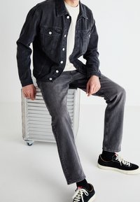 Donkerdenim jack met drukknoopsluiting over een witte blouse, gecombineerd met grijze denim jeans en zwarte sneakers met witte zolen. Zittend op een metalen krat.