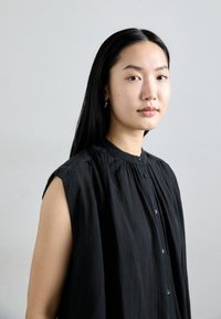 Blouse sans manches noire avec un devant boutonné, présentant un tissu texturé et un détail froncé autour de l'encolure. Le mannequin a les cheveux noirs raides.