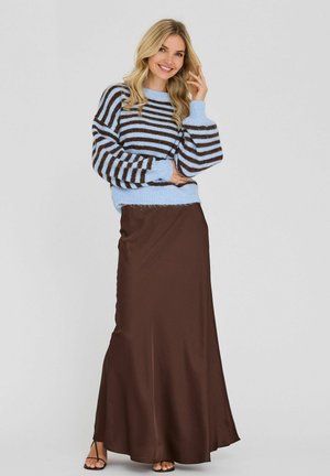 Femme souriante, portant un pull rayé bleu et marron avec des manches bouffantes et une longue jupe marron, posant devant un fond clair uni.