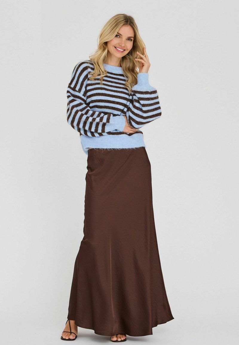 Femme souriante, portant un pull rayé bleu et marron avec des manches bouffantes et une longue jupe marron, posant devant un fond clair uni.