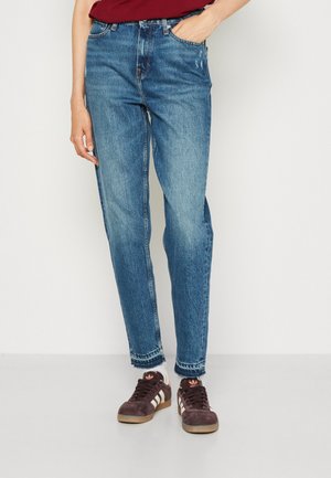 Personne portant un jean denim bleu avec des ourlets effilochés, des baskets Adidas marron avec des bandes blanches et une chemise bordeaux, debout contre un mur clair.