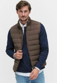 Brązowa pikowana kamizelka z wysokim kołnierzem i zamkiem błyskawicznym, w połączeniu z granatowym swetrem i jasnymi dżinsami. Charakteryzuje się gładką fakturą i równomiernym wypełnieniem.