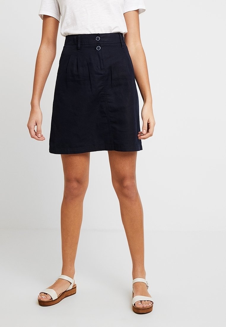 Jupe en coton bleu marine à taille haute, plissée, avec deux boutons sur le devant et un ourlet droit. Portée avec des sandales blanches à semelle en bois.