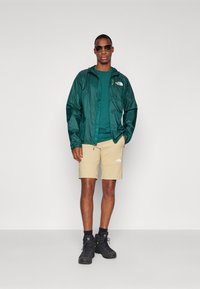 Groene lichtgewicht regenjack, teal shirt, beige shorts, zwarte laarzen en een zonnebril. Opmerkelijke logo's op het jack en de shorts. Glad textuur.