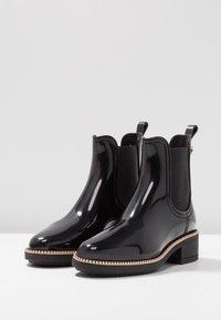 Bottines noires brillantes avec des panneaux élastiques sur les côtés, un petit talon épais et une semelle en caoutchouc texturée arborant un design en zigzag autour de la base.