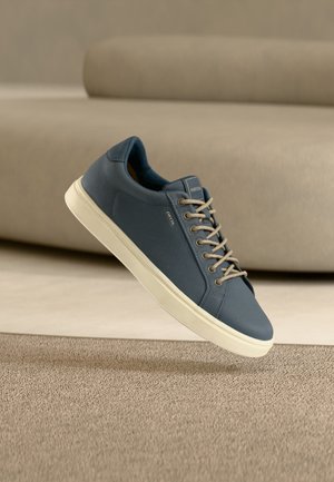 Mėlyna žema sportinė avalynė su smėlio spalvos raišteliais ir balta padu, skriejanti virš tekstūruotos smėlio spalvos kiliminės dangos prie smėlio spalvos sofos.