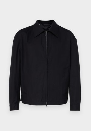 Črna jakna iz gladkega blaga, z zadrgo spredaj, s koničastim ovratnikom in dvema stranskima žepoma.