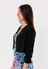 Giacca nera corta con una texture liscia e maniche a tre quarti, indossata sopra un vestito colorato e stampato con tonalità vivaci.
