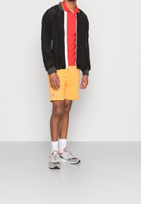 Czarna kurtka bombowca z białym zamkiem, czerwona koszulka polo, żółte szorty, szare sneakersy i białe skarpetki. Szorty mają logo.