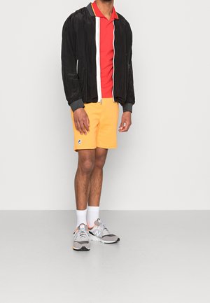 Černá bomber bunda s bílým zipem, červené polo tričko, žluté šortky, šedé tenisky a bílé ponožky. Šortky mají logo.