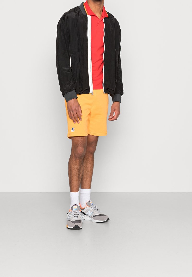 Czarna kurtka bombowca z białym zamkiem, czerwona koszulka polo, żółte szorty, szare sneakersy i białe skarpetki. Szorty mają logo.